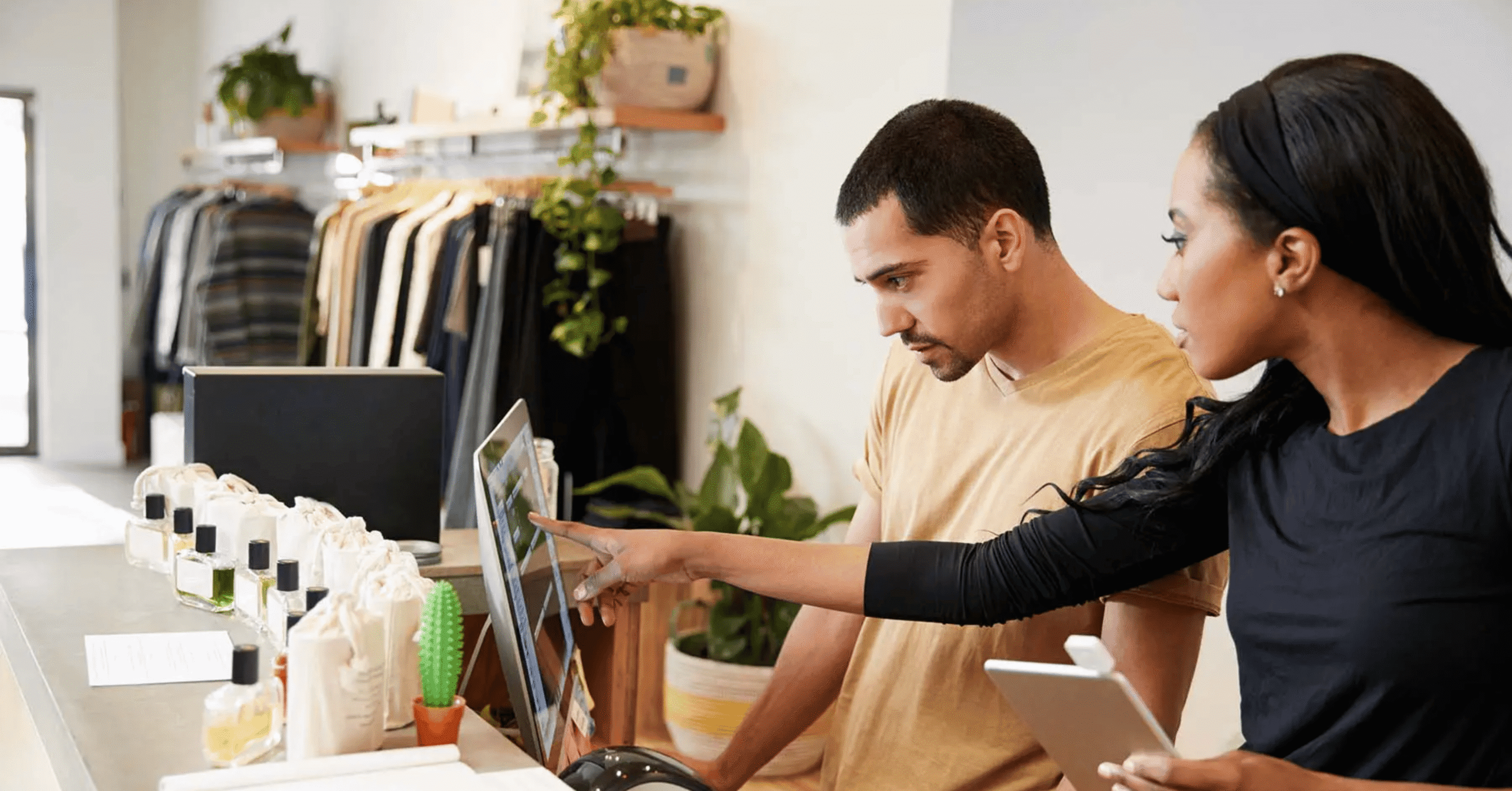 Two people stand behind a retail counter looking at a computer monitor. One person is pointing at the screen whilst the other watches. Clothing and products are displayed in the background.
